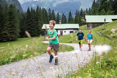 Family hiking in Gemsteltal, a boy runs laughing ahead of the group, surrounded by green nature and mountains. | © Kleinwalsertal Tourismus eGen • Photographer: Bastian Morell