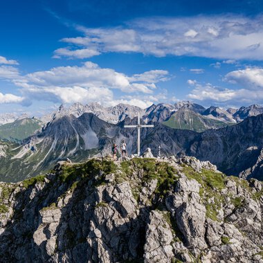 Berggipfel mit Gipfelkreuz und Wanderern, oberhalb der Alpen, bei schönem Wetter, mit blauer Himmel und Wolken. | © Kleinwalsertal Tourismus eGen • Fotograf: Bastian Morell