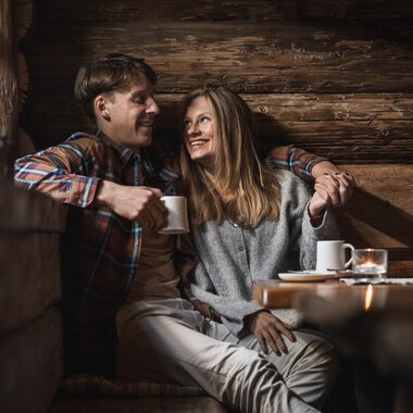 Ein Mann und eine Frau sitzen gemütlich in einer Holzhütte, lachen und trinken Kaffee, umgeben von warmem Holzinterieur. | © Kleinwalsertal Tourismus eGen • Fotograf: Hannah Bichay