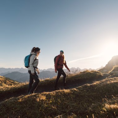 Drei Wanderer mit Rucksäcken auf schmalem Pfad in einer Berglandschaft bei Sonnenuntergang. | © Kleinwalsertal Tourismus eGen • Fotograf: Hannah Bichay