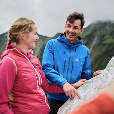 Zwei Wanderer mit Rucksack, lachend und sich unterhaltend während einer Herbstwanderung im Kleinwalsertal, mit Bergpanorama im Hintergrund. | © Kleinwalsertal Tourismus eGen • Fotograf: Oliver Farys