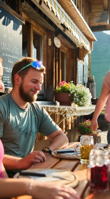 Drei Freunde genießen ein gemeinsames Essen auf einer Terrasse mit Blick auf die Berge, eine Frau steht lachend und trinkt, während im Hintergrund Blumen und Berglandschaft zu sehen sind. | © Kleinwalsertal Tourismus eGen • Fotograf: Oliver Farys
