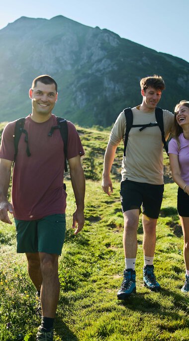 Drei junge Erwachsene wandern fröhlich auf einer grünen Wiese mit Bergen im Hintergrund bei Sonnenschein. | © Kleinwalsertal Tourismus eGen • Fotograf: Oliver Farys