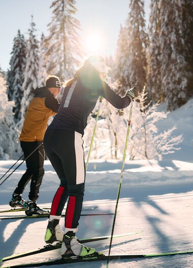 Zwei Skifahrer beim Langlaufen in verschneiter Winterlandschaft, Sonnenlicht durch Bäume, alpine Natur im Hintergrund. | © Kleinwalsertal Tourismus eGen • Fotograf: Oliver Farys