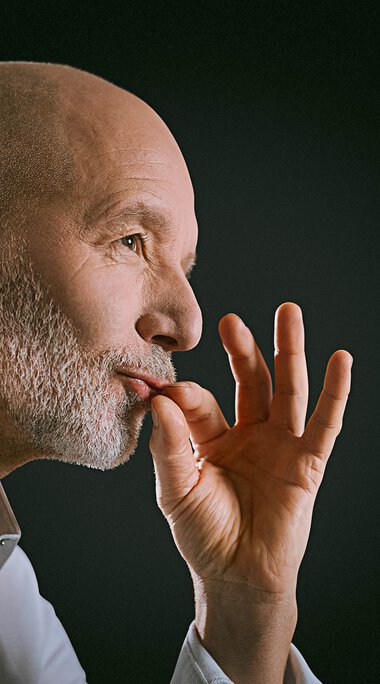 Older man in a white shirt looking into the distance with his hand near his face, against a dark background. | © Kleinwalsertal Tourismus eGen • Photographer: Oliver Farys