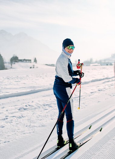 Skifahrer auf der Langlaufloipe im Schnee, umgeben von Bergen und einer Winterlandschaft, mit einer Person im Vordergrund, die einen Skistock hält. | © Kleinwalsertal Tourismus eGen • Fotograf: Oliver Farys