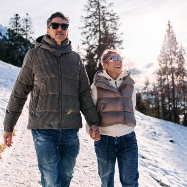 Ein Mann und eine Frau wandern im winterlichen Schnee, umgeben von Tannenbäumen, bei Sonnenschein, tragen warme Kleidung. | © Kleinwalsertal Tourismus eGen • Fotograf: Oliver Farys