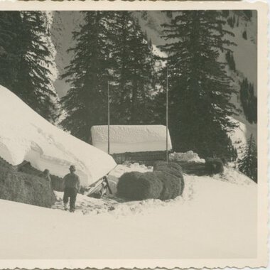 Historische Schwarz-Weiß-Aufnahme eines Hauses in verschneiter Landschaft, umgeben von Tannenbäumen, mit einem Menschen davor. | © Kleinwalsertal Tourismus eGen • Stefan Heim