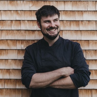 A smiling chef in a black chef jacket standing in front of a wooden shingle wall, arms crossed. | ©  Kleinwalsertal Tourismus eGen • Naturhotel Chesa Valisa