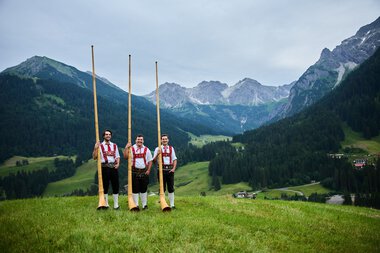 Drei Männer in Trachten stehen in einer grünen Wiese mit Alphörnern, umgeben von Bergen und einer bewaldeten Talregion.